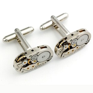 Pode incluir: Um par de bot&otilde;es de punho de prata com um design de movimento de rel&oacute;gio vintage. Os bot&otilde;es de punho apresentam uma base de prata com detalhes dourados e o texto "ELGIN U.S.A. 15 JEWELS 3675458" na face.