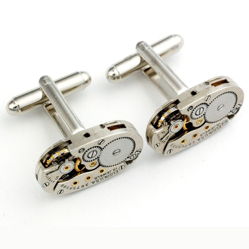 Pode incluir: Um par de bot&otilde;es de punho de prata com um design de movimento de rel&oacute;gio vintage. Os bot&otilde;es de punho apresentam uma base de prata com detalhes dourados e o texto "ELGIN U.S.A. 15 JEWELS 3675458" na face.