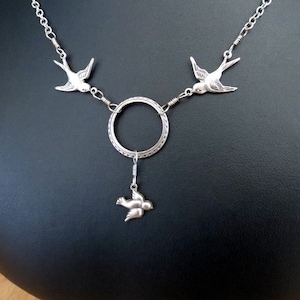 May include: Silver necklace with three bird charms. The necklace features a circle pendant with a dangling bird charm.