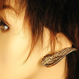 May include: A gold-toned wing-shaped earring with a detailed design. The earring is worn on the left ear of a person with brown hair.