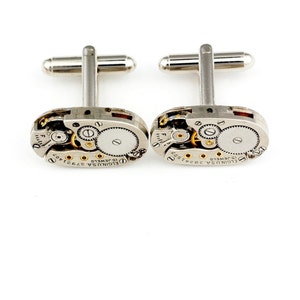 Pode incluir: Um par de bot&otilde;es de punho de prata com um design de movimento de rel&oacute;gio vintage. Os bot&otilde;es de punho apresentam um corpo de metal prateado com engrenagens expostas e uma inscri&ccedil;&atilde;o dourada que diz "ELGIN USA 15 JEWELS 3792402".