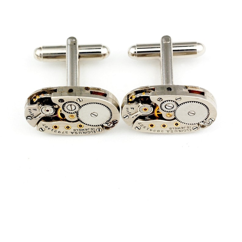 Pode incluir: Um par de bot&otilde;es de punho de prata com um design de movimento de rel&oacute;gio vintage. Os bot&otilde;es de punho apresentam um corpo de metal prateado com engrenagens expostas e uma inscri&ccedil;&atilde;o dourada que diz "ELGIN USA 15 JEWELS 3792402".