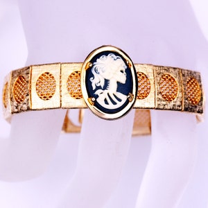 May include: A gold tone bracelet with a black and white cameo of a woman's profile. The bracelet has a mesh design with circular cutouts.