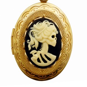 May include: A gold locket with a black and white cameo of a skull with a feminine hairstyle. The cameo is set in a gold oval frame with a detailed design.