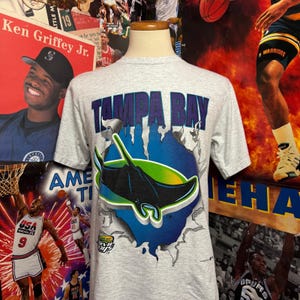 May include: A gray t-shirt featuring the text "TAMPA BAY" in blue and a graphic of a manta ray. The design includes blue and green accents, with a torn-paper effect. The shirt is displayed on a mannequin.