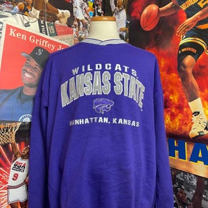 May include: A purple sweatshirt with the words "Wildcats Kansas State" in silver lettering. Below the text is the city name "Manhattan, Kansas". The sweatshirt has a white and gray striped collar.