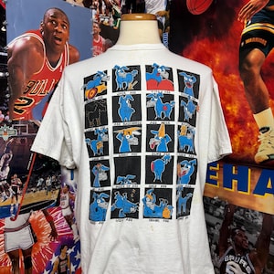 May include: White t-shirt featuring a grid of blue cartoon dog illustrations with various expressions and phrases like "ASS BACKWARDS" and "DUMB ASS". The shirt has a crew neck and short sleeves.