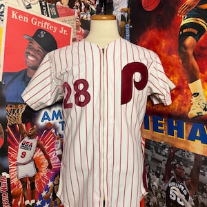 May include: A vintage baseball jersey with red vertical stripes on a white background. The jersey features the number 28 and a large maroon "P" on the front. It has a full zipper and short sleeves. The jersey is displayed on a mannequin.