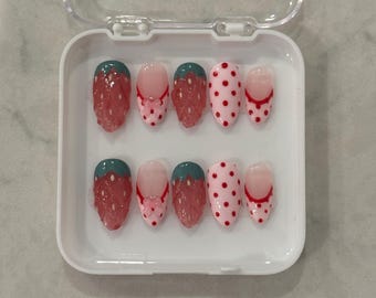 Puede incluir: Un juego de diez uñas postizas en un contenedor de plástico transparente. Las uñas están decoradas con un diseño de fresa, lunares rojos y un lazo.