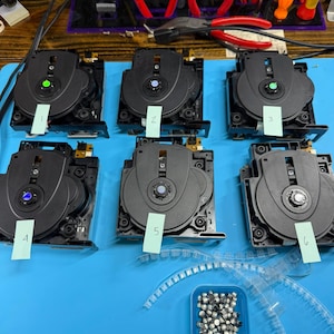 May include: Six black optical disc drive mechanisms arranged in two rows, each labeled with a numbered green paper tag from one to six. A small blue container filled with tiny metal components sits below the mechanisms on a blue mat.