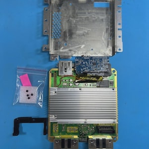 May include: Disassembled video game console components on a blue surface. Includes a silver metal casing, circuit boards with various chips and connectors, and small plastic bags with components. The components are arranged for repair or modification.