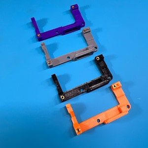 May include: Four 3D-printed L-shaped brackets in purple, gray, black, and orange, each with small gold-colored fasteners. The brackets are arranged diagonally on a blue surface, showcasing their unique design and construction.