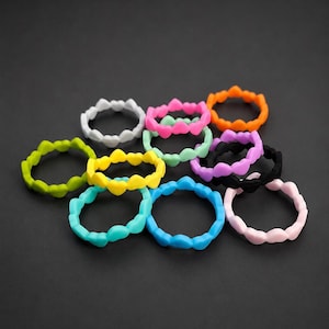 Op de afbeelding: Een verzameling van elf ronde ringen, elk met een herhalend hartvormig patroon langs de band. De ringen zijn uitgevoerd in verschillende effen, levendige kleuren, waaronder wit, roze, oranje, limoengroen, geel, zwart, lichtblauw en talingblauw.