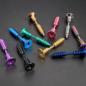 Könnte beinhalten: Eine Sammlung von schraubenförmigen Ohrringen in verschiedenen metallischen und leuchtenden Farben, darunter Gold, Silber, Schwarz, Blau, Grün, Lila und Pink. Jeder Ohrring hat einen Gewindestift, einen flachen Kopf mit Kreuzschlitz und ein spitzes Ende.