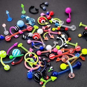 May include: A collection of colorful body jewelry, including curved barbells, straight barbells, and captive bead rings. The jewelry features various designs, such as striped balls, arrow shapes, and clear orbs. Colors include pink, blue, green, red, and black.