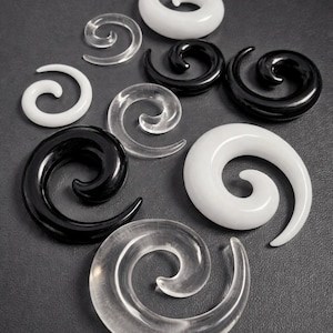 May include: An assortment of spiral ear gauges in clear, black, and white. The gauges vary in size and are displayed on a dark grey surface. The image highlights the different designs and materials used in the ear jewellery.