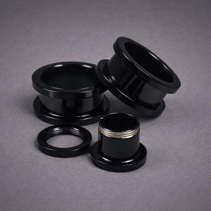 Peut inclure: Plusieurs écarteurs d'oreille noirs de différentes tailles sont disposés sur une surface grise. Les écarteurs ont une finition lisse et brillante. L'un d'eux a un anneau argenté au centre. Conçus pour les piercings d'oreille étirés.