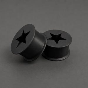 Peut inclure: Deux écarteurs d'oreille gris foncé avec une découpe en forme d'étoile au centre. Les écarteurs sont cylindriques avec un bord évasé et reposent sur une surface grise. La découpe en étoile est centrée sur la face de chaque écarteur.