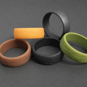 May include: Five textured rings in shades of brown, orange, black, grey, and green. The rings have a wood-grain effect and are made of a solid material. They are arranged on a dark grey surface.