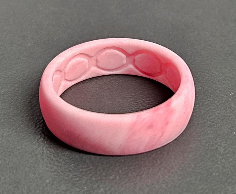 May include: A pink silicone ring with a marbled effect. The inside of the ring features a series of oval-shaped cutouts. The ring is a solid, rounded band, suitable for everyday wear.