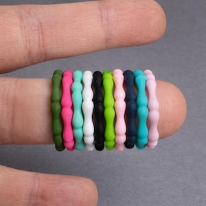 Op de afbeelding: Een verzameling kleurrijke siliconen ringen in verschillende tinten, waaronder groen, roze, wit, zwart en blauw. De ringen zijn op een vinger gestapeld en laten hun flexibele en getextureerde ontwerp zien. De achtergrond is effen grijs.