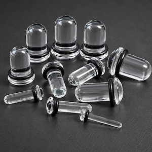 Op de afbeelding: Een collectie transparante glazen oorplugs in verschillende maten, elk met een afgeronde voorkant en een glad cilindrisch lichaam. Elk sieraad is voorzien van een dunne zwarte rubberen O-ring om het op zijn plaats te houden.