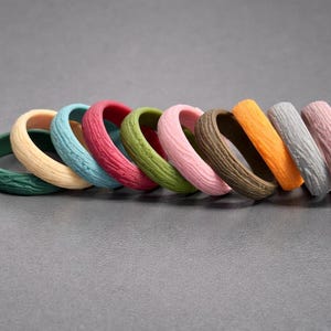 May include: Nine rings in a row, each with a textured, wood-like finish. The rings are in various colours: green, beige, blue, red, green, pink, brown, orange, grey, and pink. The rings are made of a durable material and are suitable for everyday wear.