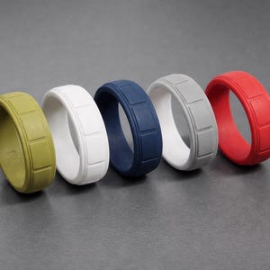 May include: Five silicone rings in olive green, white, navy blue, grey, and red. Each ring has a rectangular pattern around the band. The rings are displayed on a grey surface.