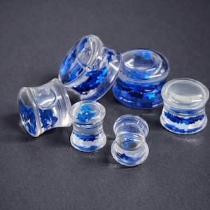May include: Clear acrylic ear gauges with blue star-shaped glitter. The plugs are in various sizes and shapes, showcasing a translucent design with embedded blue accents. The jewelry is displayed on a dark gray surface.