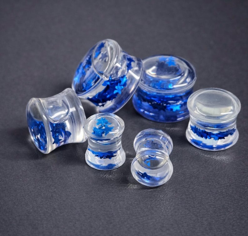 May include: Clear acrylic ear gauges with blue star-shaped glitter. The plugs are in various sizes and shapes, showcasing a translucent design with embedded blue accents. The jewelry is displayed on a dark gray surface.