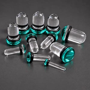 May include: A collection of clear glass ear plugs and tapers featuring vibrant teal-coloured ends and thin black rubber O-rings. The set includes various sizes of cylindrical body jewellery with rounded tips, arranged in a scattered formation.