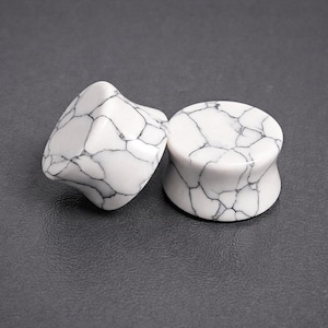 May include: Two white and black marbled stone ear plugs. One plug is rounded and the other is a more organic shape. The plugs are made of a white stone with black veining throughout. The plugs are designed to be worn in stretched ear piercings.