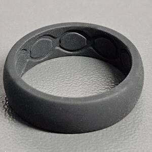 May include: A dark gray silicone ring with a textured surface. The ring features three oval-shaped designs on the top. The ring is a solid color and appears to be flexible.