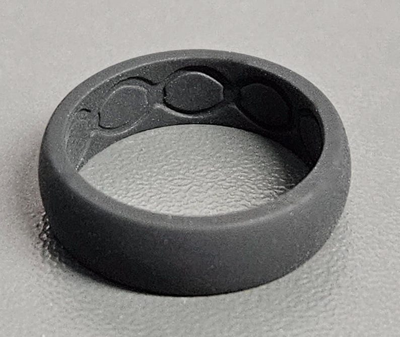 May include: A dark gray silicone ring with a textured surface. The ring features three oval-shaped designs on the top. The ring is a solid color and appears to be flexible.