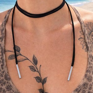 May include: A black choker necklace with a double-strand design. The necklace has long, thin black cords with silver-coloured metal tips. The necklace is worn over a patterned top.
