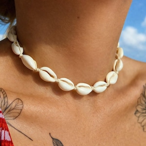 May include: A cream-coloured cowrie shell necklace. The shells are strung together with a beige cord, creating a choker style. The shells are oval-shaped and have a smooth, glossy texture. The necklace is worn around the neck.