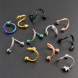 May include: A collection of body jewelry including spiral, circular, and curved barbell pieces in various finishes such as gold, silver, rose gold, black, and iridescent rainbow. The items feature ball, spike, and star-shaped end attachments.