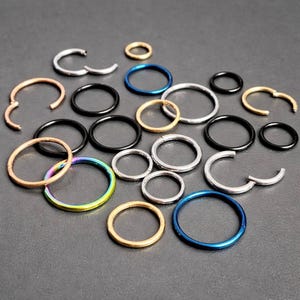 May include: A selection of hoop earrings in various sizes and colours, including silver, gold, black, and blue. Some hoops are open, while others are closed. The earrings are arranged on a dark grey surface.
