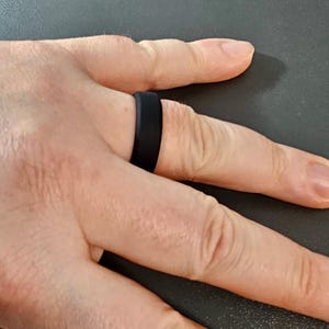 Op de afbeelding: Een zwarte siliconen ring wordt aan een vinger gedragen. De ring is een effen band met een matte afwerking. De achtergrond is een donkergrijze ondergrond.