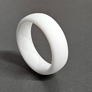 May include: A white silicone ring with a smooth exterior and a patterned interior. The ring is a solid, opaque white color and appears to be flexible. The design features a series of circular indentations on the inside.