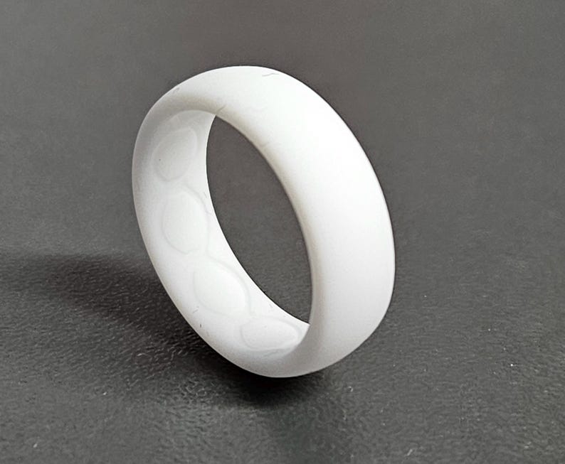 May include: A white silicone ring with a smooth exterior and a patterned interior. The ring is a solid, opaque white color and appears to be flexible. The design features a series of circular indentations on the inside.