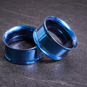 Op de afbeelding: Twee blauwe, metalen oortunnels. De tunnels zijn cirkelvormig met een uitlopende rand en een glanzend, reflecterend oppervlak. Ze liggen op een donkere, getextureerde ondergrond.