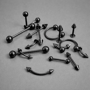 May include: A selection of black body jewellery, including barbells, curved barbells, and circular barbells. Some pieces have ball ends, while others feature cone-shaped spikes. The jewellery is displayed on a grey surface.