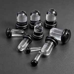 May include: A collection of clear glass ear plugs with rounded ends and black rubber O-rings. The set includes various sizes of these cylindrical body jewellery pieces, each featuring a smooth, transparent body and a solid black ring at the base.