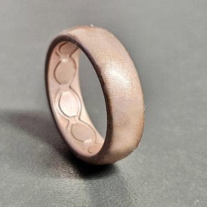 May include: A rose gold silicone ring with a textured surface. The inside of the ring features a series of connected oval shapes. The ring is designed with a matte finish and a slightly darker edge.