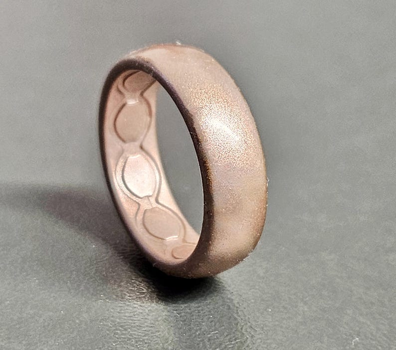 May include: A rose gold silicone ring with a textured surface. The inside of the ring features a series of connected oval shapes. The ring is designed with a matte finish and a slightly darker edge.