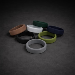 Könnte beinhalten: Eine Sammlung von Silikonringen in verschiedenen Farben, darunter Braun, Schwarz, Grau, Weiß, Grün und Marineblau. Die Ringe sind auf einer dunkelgrauen Oberfläche angeordnet und zeigen ihre glatte, matte Oberfläche. Diese Ringe sind für den täglichen Gebrauch konzipiert.