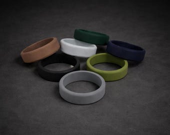 May include: A selection of silicone rings in diverse colours, such as brown, black, grey, white, green, and navy blue. The rings are displayed on a dark grey surface, highlighting their smooth, matte finish. These rings are designed for everyday wear.