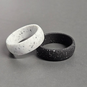 May include: Two speckled silicone rings. One is white with black speckles, the other black with white speckles. The rings are round and have a smooth, matte finish. They are likely used as wedding bands or fashion accessories.