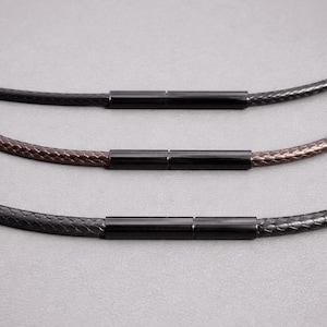 May include: Three braided leather necklaces in black, brown, and dark grey, each with a black cylindrical clasp. The necklaces are displayed against a neutral grey background, showcasing the texture and color variations of the leather.